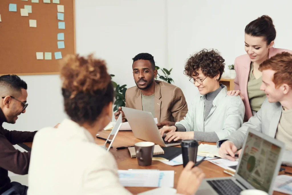 A group of coworkers smiling and talking together while sitting at a wooden meeting table with laptops and coffee.