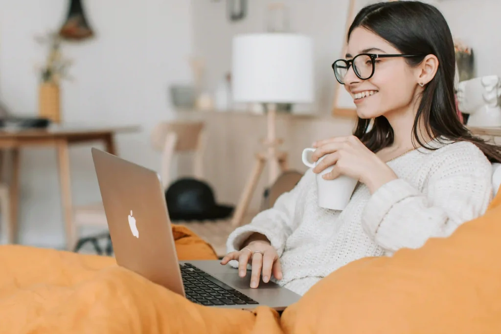 A woman wearing glasses relaxes comfortably on a yellow beanbag chair while using her laptop and holding a coffee mug.