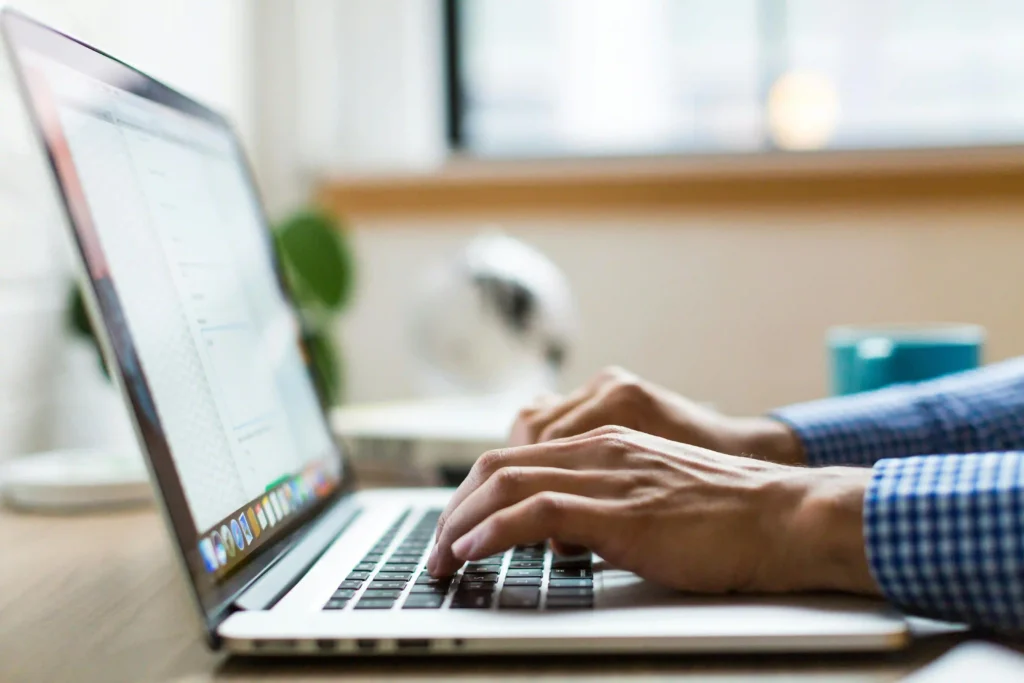 A side profile close-up of a person's hands typing on a laptop keyboard in a brightly lit office.