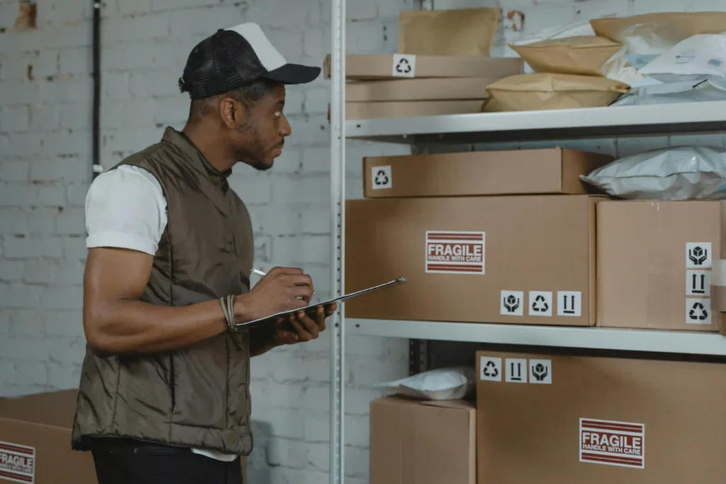A warehouse worker wearing a cap and safety vest checks inventory on a clipboard next to stacked cardboard boxes.